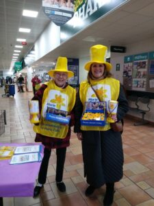 Soroptimist member and Marie Curie volunteer in tabards and hats collecting donations.