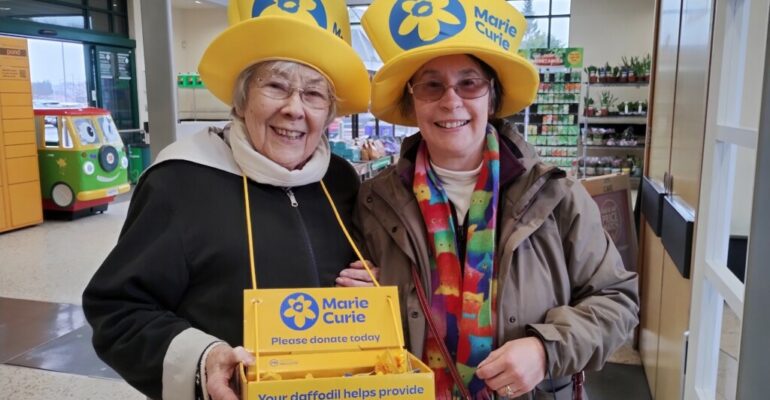 Two smiling women wearing yellow Marie Curie hats. Both hold donation boxes.
