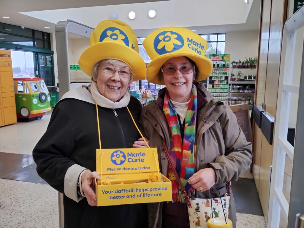 Two smiling women wearing yellow Marie Curie hats. Both hold donation boxes.