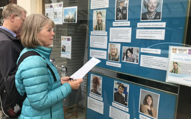 woman in turquoise jacket holding a sheet of paper looking at display in window