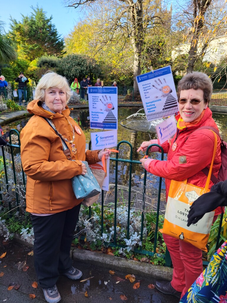 Valerie is smiling, dressed in a orange jacket, holding a sign. Lydia smiles to the camera, while holding her sign under one arm, as she ties a ribbon. She carries an orange bag.