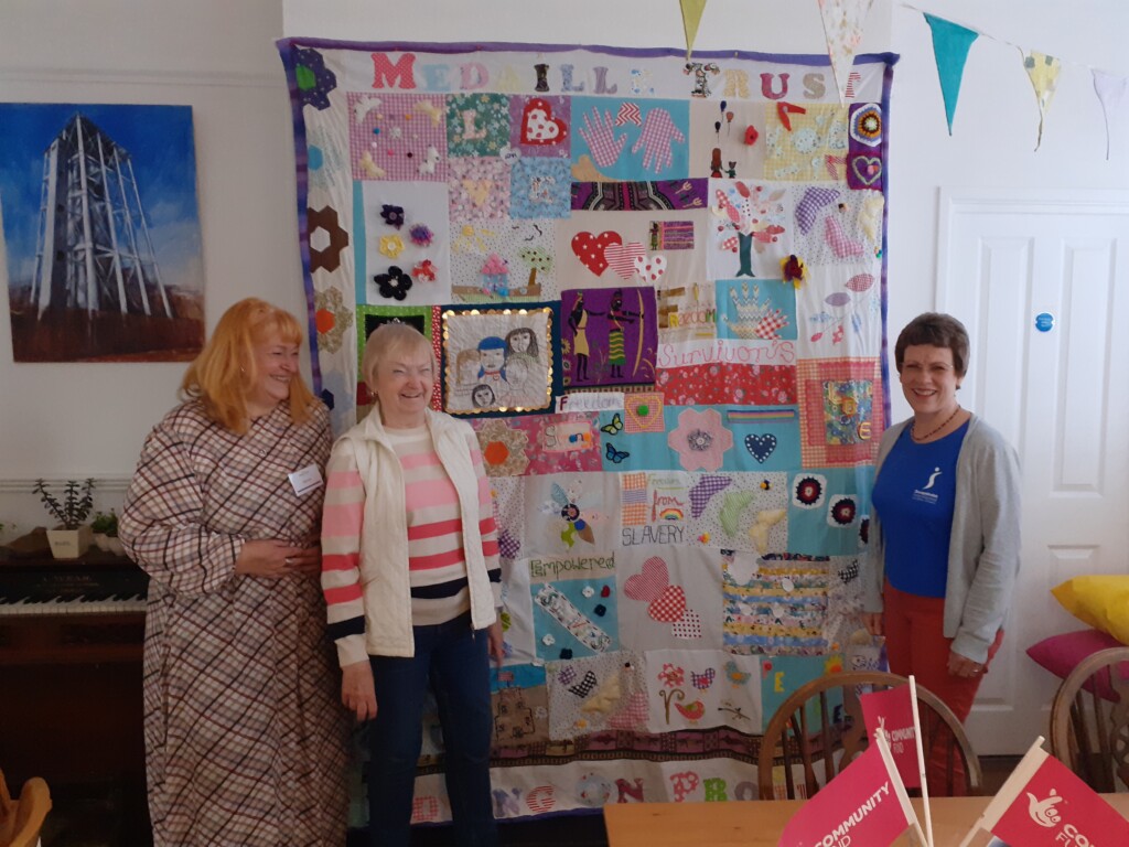 Donna and Jackie stand to the left of the stupendous quilt made by the clients of the Moving on Project. Lydia stands to the right, wearing a blue Soroptimist t-shirt